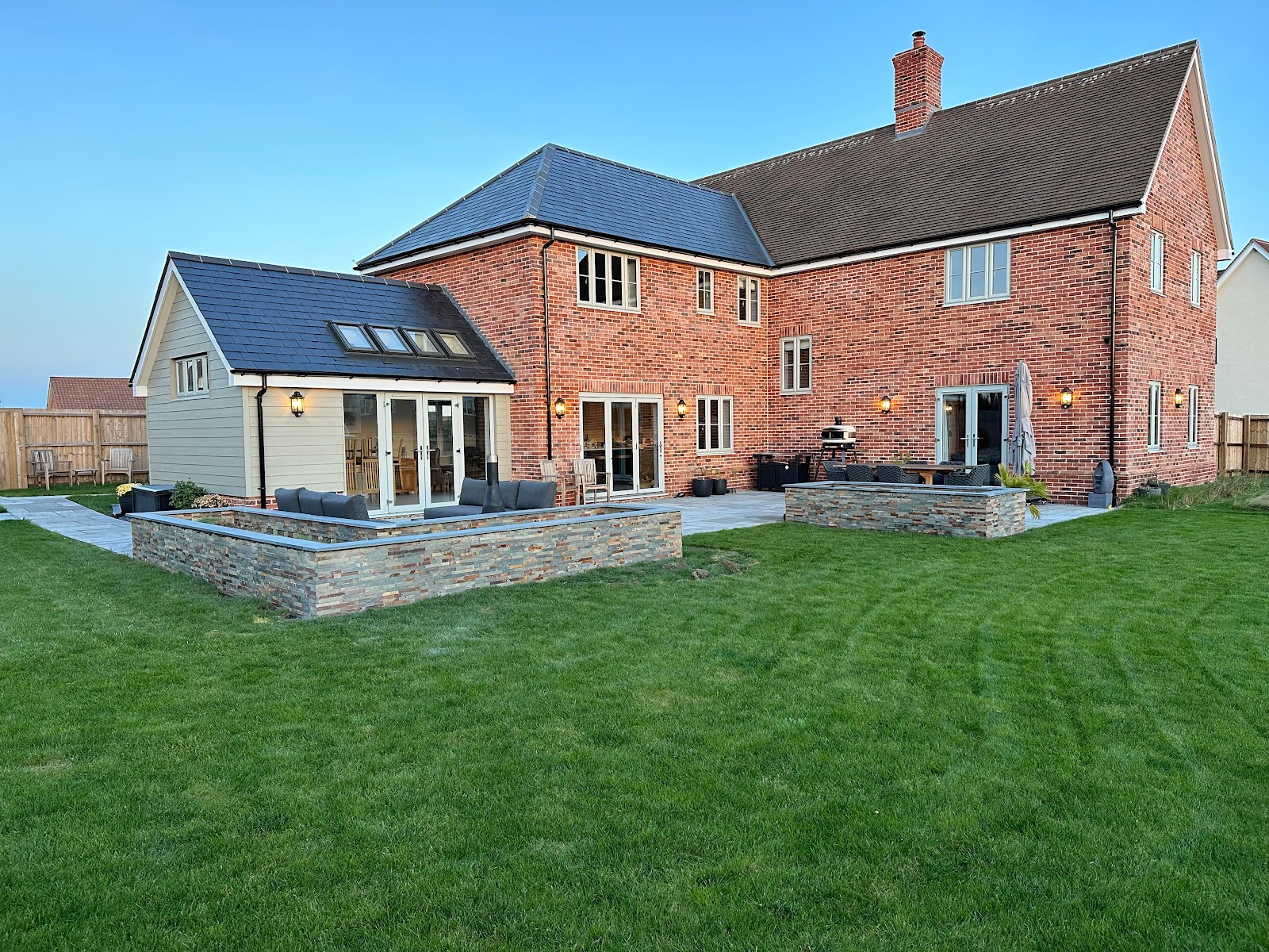 Lawn, paved terrace and stone clad raised beds behind a large red brick house, laid by A&J Fox Paving